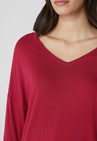 Rotes V-Ausschnitt-Shirt mit langen Ärmeln, aus weichem Stoff, mit lockerer Passform und glatter Textur, das bis zur oberen Brust reicht.