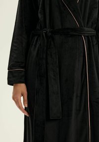 Czarna, welurowa szata o gładkiej fakturze, z zawiązywanym paskiem w talii i kontrastującym różowym obszyciem przy kołnierzu i mankietach.