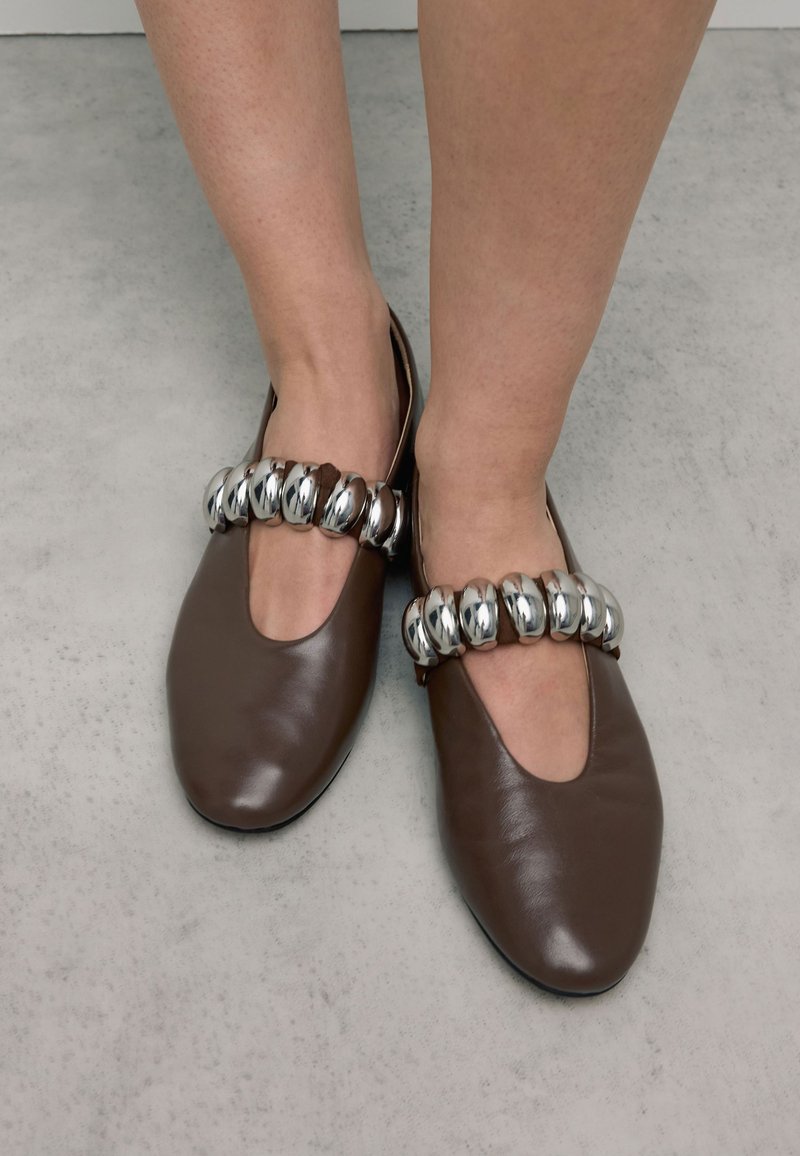 Des pieds portant des chaussures en cuir marron à enfiler avec une large bride en perles argentées sur le dessus, se tenant sur un sol gris.