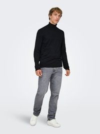 Svart polotröja i figurnära design kombinerad med ljusgrå jeans och vita sneakers, stående mot en minimalistisk vit bakgrund.
