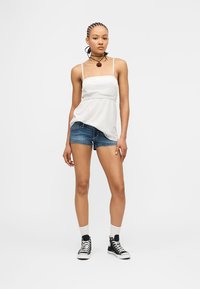 Γυναίκα με πλεγμένα μαλλιά που φοράει λευκό τοπ με λεπτές τιράντες, σορτς από τζιν, μαύρα αθλητικά παπούτσια ψηλά και κολιέ με κρεμαστό λουλούδι.