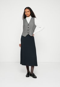 Gray, fitted vest with four buttons over a white turtleneck, paired with a long, pleated navy skirt and black shoes.