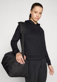 Svart duffelbag med strukturert stoff, avtagbar skulderreim og glidelås. Kombinert med en matchende svart hettegenser og leggins.