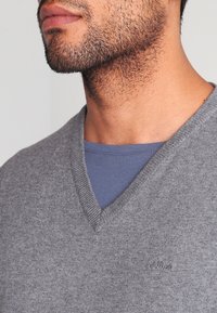 Pull gris à col en V en maille douce, porté sur une chemise bleue. Présente un logo discret brodé sur le pull. Tissu texturé.