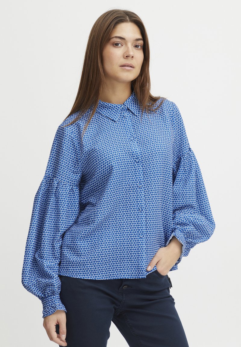 Blusa blu a fantasia con design a bottoni, caratterizzata da un colletto e maniche lunghe e voluminose. Realizzata in un tessuto testurizzato.