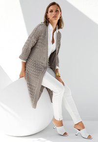 Leichter, gestrickter Cardigan in Taupe mit offenem Front, kombiniert mit weißen Slim-Fit-Hosen und weißen offenen Mules mit kleinem Absatz.