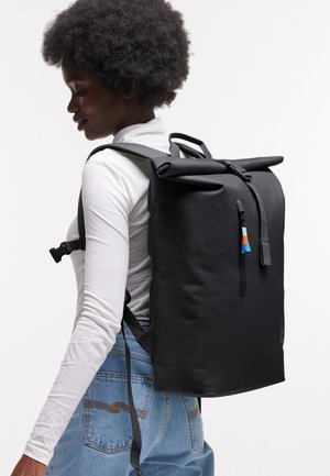 Giovane con capelli ricci che indossa una maglia bianca a maniche lunghe, jeans di denim e uno zaino nero grande con chiusura a rotolo.