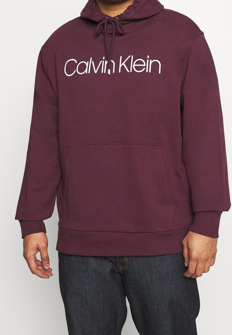 Sudadera burdeos con capucha ajustable, gran bolsillo frontal y logo blanco de "Calvin Klein". Hecha de material suave con puños acanalados.