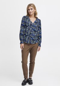 Blusa a stampa floreale blu e bianca su sfondo nero, con maniche lunghe e scollatura a V, abbinata a pantaloni slim fit marroni e scarpe chunky.