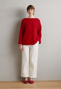 Rdeča pletena sv sweater z širokim izrezom in dolgimi rokavi, v kombinaciji z belimi širokimi hlačami in rdečimi ravnimi čevlji. Vidna tekstura tkanine.