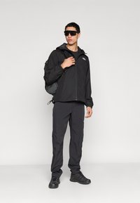 Μαύρο αδιάβροχο μπουφάν με κουκούλα, σετ με μαύρο παντελόνι και ανθεκτικά μαύρα παπούτσια. Περιλαμβάνει σακίδιο πλάτης και γυαλιά ηλίου.