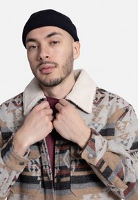 Wollen jas met een patroon in aardetinten, voorzien van een crèmekleurige sherpa-kraag, frontknopen en een bordeauxrode overhemd eronder.