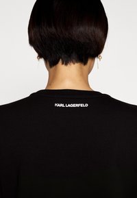 Persona con cabello corto y oscuro que lleva una camiseta negra con el texto "KARL LAGERFELD" en la parte posterior cerca del cuello, y aretes de perlas colgantes.