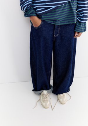 Személy túlméretezett sötétkék farmernadrágot, csíkos hosszú ujjú inget és fehér, kikötetlen cipőfűzőjű tornacipőt visel, fehér háttér előtt áll.