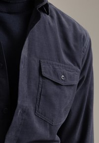 Camisa de pana azul marino con cuello abotonado, que presenta un bolsillo en el pecho y una textura suave, parcialmente superpuesta sobre un cuello de tortuga oscuro.