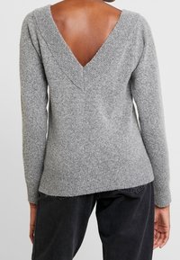 Pull en laine gris avec un dos en V profond, des poignets et un ourlet côtelés, et une texture douce. Convient à une tenue décontractée, à associer avec un pantalon sombre.