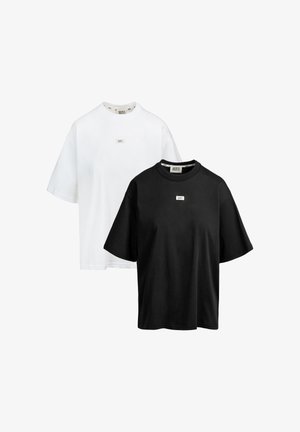 Dwa oversize T-shirty z krótkim rękawem, jeden biały i jeden czarny, każdy z małą prostokątną naszywką z logo na piersi oraz metką marki na kołnierzu.