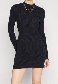 Svart ribbad långärmad klänning med en åtsittande silhuett, rund halsringning och strukturerad tyg. Lager av guldhalsband som accessoarer.