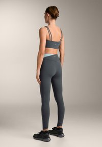 Harmaa urheilurintaliivi, jossa on ohuet hihnat, ja siihen sopivat korkeavyötäröiset legginssit. Kangas näyttää sileältä ja hiukan kiiltävältä. Tumma jalkineet täydentävät asun.