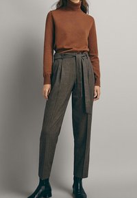 Femme portant un pull à col roulé marron, un pantalon taille haute à carreaux gris avec une ceinture, et des bottines noires, sur un fond uni.