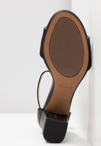 Semelle d'une sandale noire Clarks avec un talon bloc bas, une semelle intérieure beige et une fine lanière au niveau de la cheville, inclinée contre un fond d'angle blanc.