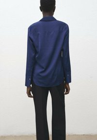 Navy blue long-sleeve shirt with a relaxed fit, back yoke detail, and subtle collar. Paired with black wide-legged trousers.