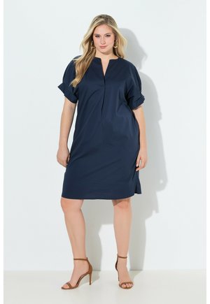 Woman wearing a navy blue knee-length dress with rolled sleeves and brown high-heeled sandals, standing against a white background.