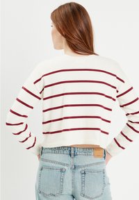 Weißer verkürzter Pullover mit burgunderfarbenen horizontalen Streifen, weiche Stricktextur. Kombiniert mit hochgeschnittenen hellblauen Jeans.