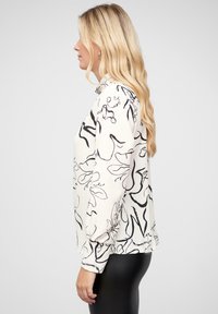 Blouse à manches longues en crème avec des motifs abstraits noirs, dotée de poignets et d'une coupe décontractée. Tissu lisse avec une texture légère.