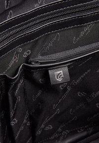 Schwarz gefütterte Stofftasche mit mehreren Reißverschlusstaschen, glatter Oberfläche und einem Logotag, der in dunklen Tönen auf einem gemusterten Innenfutter gedruckt ist.