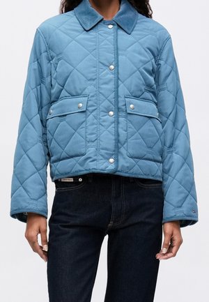 Veste matelassée bleue avec boutons-pression et deux poches avant à rabat portée avec un jean foncé, vue des épaules aux hanches.
