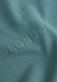 Tamsiai žalia medvilnė su lygiu paviršiumi, kuriame centre įspaustas žodis "LONDON" tamsesnėje žalioje spalvoje.