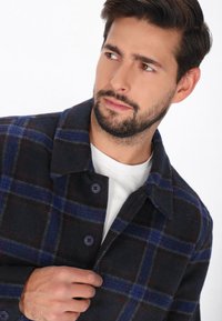 Geklede wollen jas met een geblokt patroon in donkerblauw en grijs, met rode accenten. Opvallende knopen en een klassieke kraagdetails verbeteren het ontwerp.