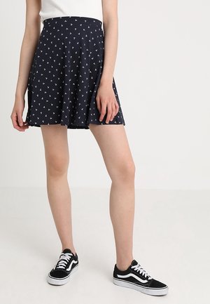 Kvinde iført navyblå nederdel med hvidt ankerprint, hvid ærmeløs top og sort-hvide snørestøvler, stående på hvidt gulv.