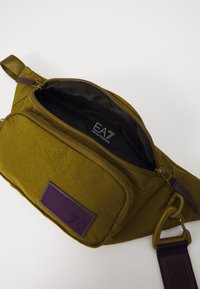 Olive green nylon bag with a textured finish, featuring a purple accent patch, zippered pocket, and adjustable strap with a clip closure.