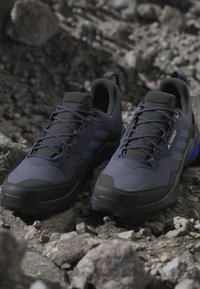 Tmavo šedé športové topánky s čiernymi akcentmi, štruktúrovaný zvršok, modré šnúrky, zosilnená špička a logo s tromi pruhmi na boku.
