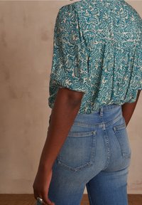 Blusa a maniche corte in verde petrolio con un motivo paisley crema, abbinata a jeans blu a vita alta. Il tessuto appare leggero e con una trama liscia.