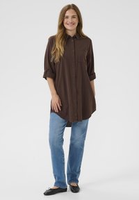 Mlada ženska z dolgimi lasmi, oblečena v temno rjavo srajco z gumbi, modre kavbojke in črne baletne copate, nasmejana in stoji.