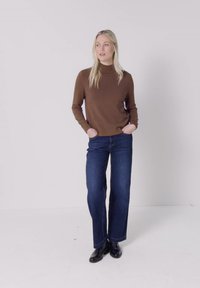 Femme aux longs cheveux blonds portant un haut marron à manches longues, un jean bleu à jambes larges et des chaussures noires, debout les mains dans les poches.