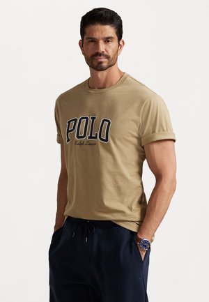 Beige bomulds T-shirt med rullede ærmer, prydet med en fremtrædende "POLO" logo i marineblå, parret med marineblå joggerbukser og et ur på håndleddet.