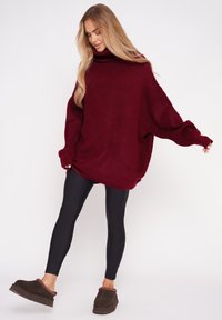 Burgundowa, oversize'owa sweterka z wysokim kołnierzem, w połączeniu z czarnymi legginsami i brązowymi butami typu slip-on z dekoracyjnym akcentem.