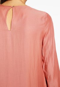 Blusa color rosa con una texture setosa, dotata di scollatura rotonda e dettaglio a chiave sul retro con un piccolo bottone dorato.