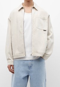 Lys beige lynlåsjakke med krave, brystlomme og to sidelommer; parret med lyseblå denimbukser, båret over en hvid tanktop.