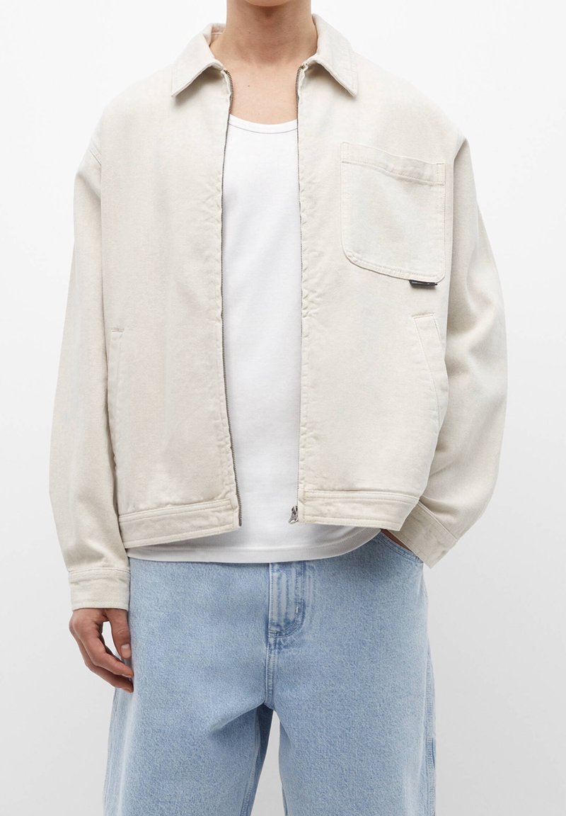 Lys beige lynlåsjakke med krave, brystlomme og to sidelommer; parret med lyseblå denimbukser, båret over en hvid tanktop.