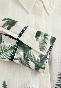 Korte mouw blouse in lichtbeige stof met groene palmbladafdruk. Heeft een puntige kraag en knoopdetails op de mouwen. Geweven stof.