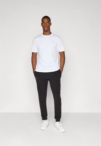 Witte t-shirt met korte mouwen en zwarte joggerbroeken. Het shirt is simpel, en de joggers hebben een subtiele logo. Schoenen zijn witte sneakers.