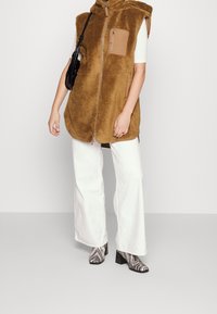 Braun, flauschige ärmellose Weste mit einem Reißverschluss und einer Brusttasche, kombiniert mit weißen weiten Hosen und schwarz-weißen gemusterten Stiefeletten mit Absatz.