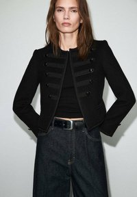 Czarna krótka kurtka z guzikiem w stylu wojskowym, z wyraźnie zarysowanymi ramionami i dopasowanym krojem. Noszona na czarnym topie i zestawiona z ciemnymi dżinsami.