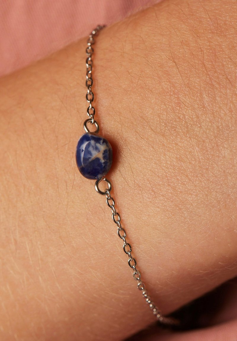 Zilveren schakelarmband met een centrale blauwe steen met witte patronen, een gladde structuur en ovale vorm, rustend op een pols.