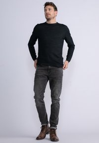 Pullover in maglia scura texturizzata con maniche lunghe, abbinato a jeans grigi slim fit e stivali stringati marroni, su uno sfondo neutro.